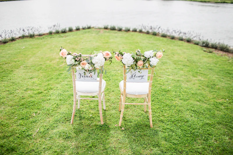 Two wooden sweetheart chairs trimmed with roses and 'Forever' and 'Always' signs on a lawn by a pond