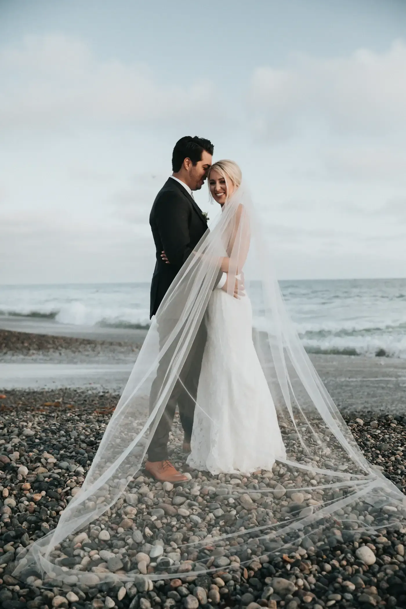 Bride and groom at the water's edge on a San Diego beach, the bride's long veil sweeping across the pebbles at dusk