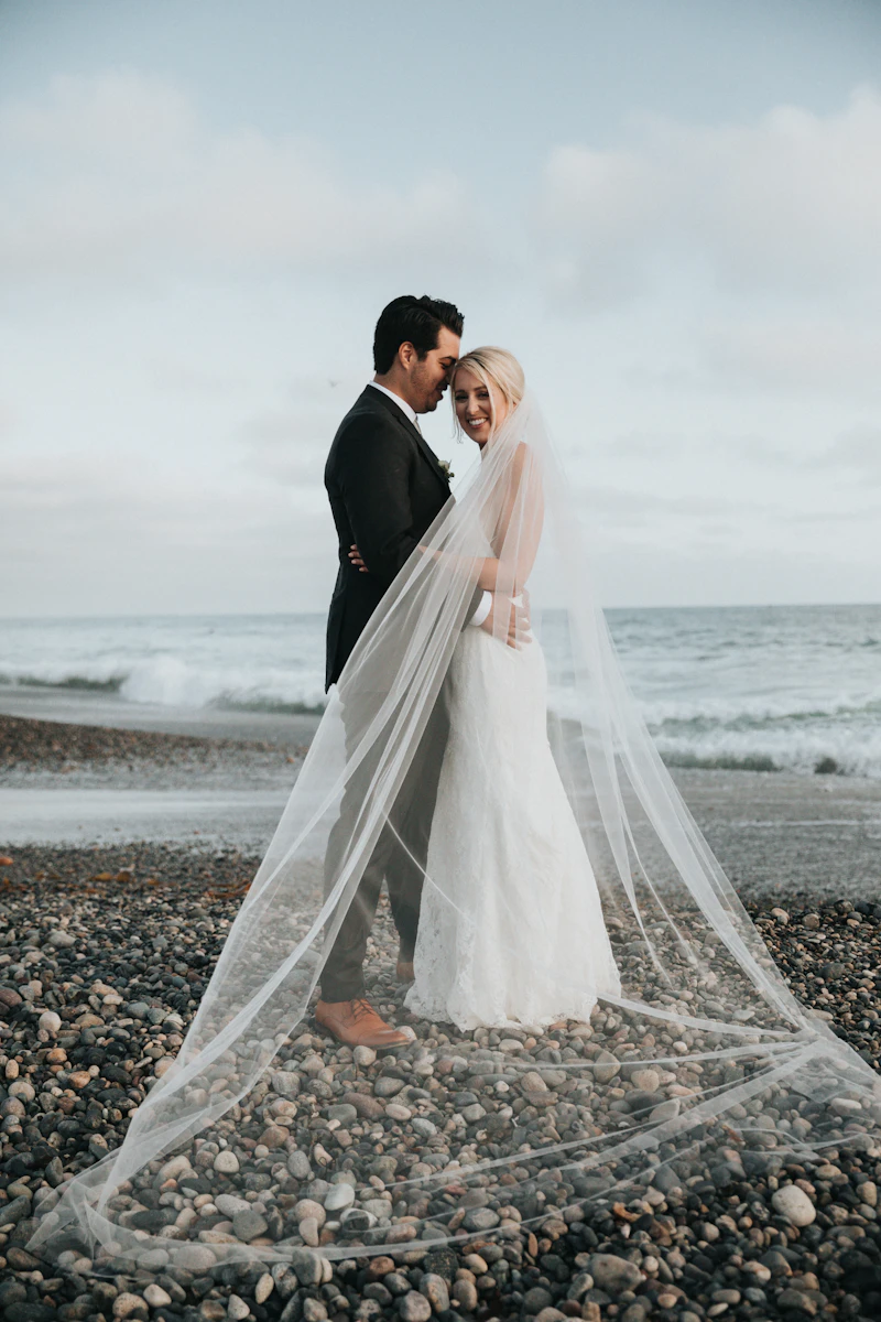 Couple embracing on a rocky Pacific coast beach, bride's cathedral veil sweeping in the wind