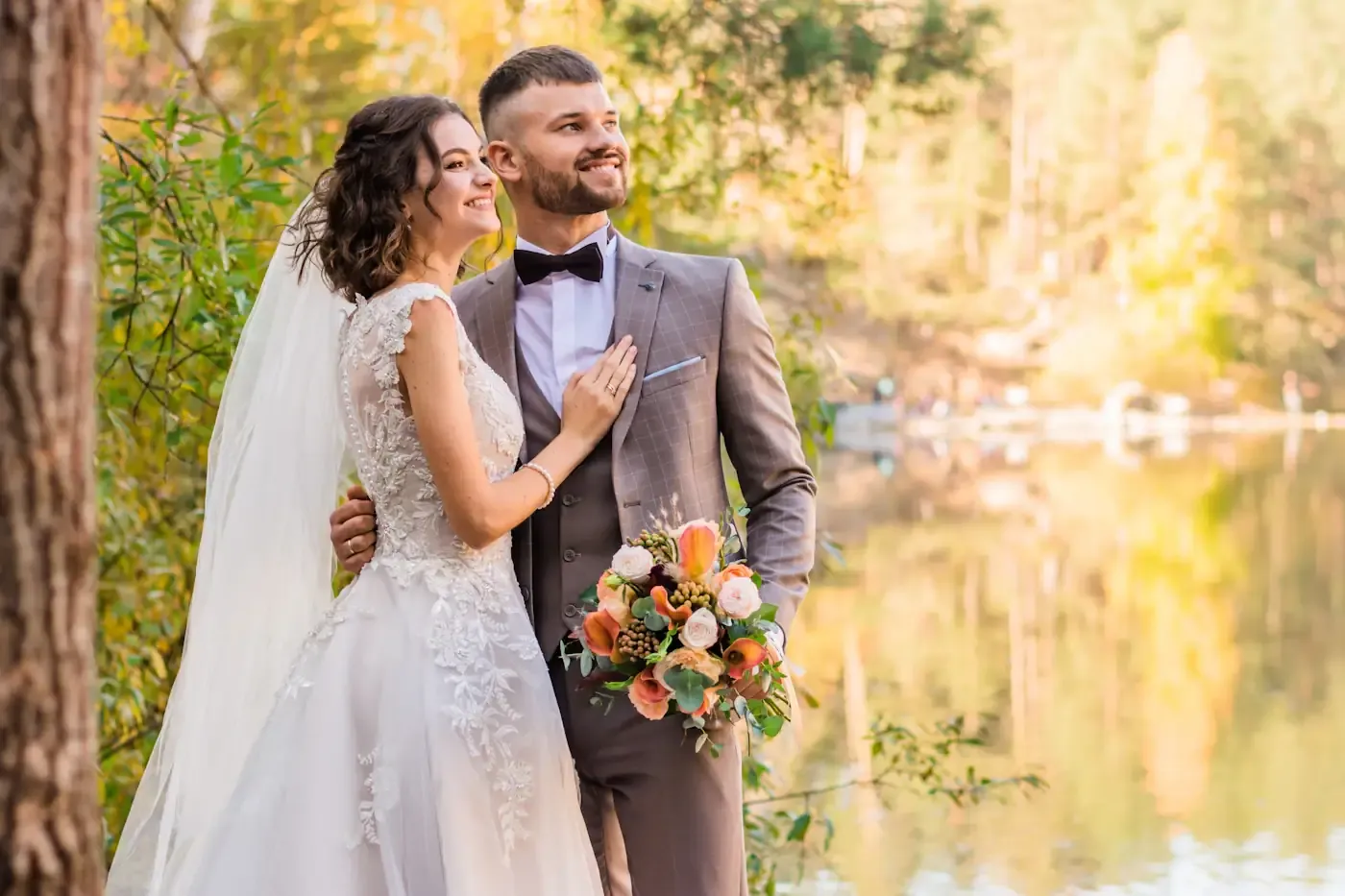 Couple embracing beside still water as the light drops, bride holding a peach bouquet