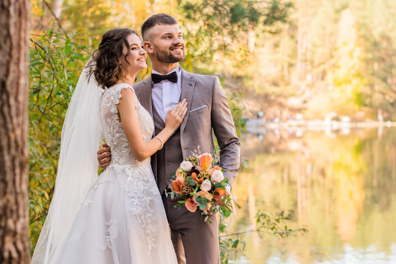Couple in elegant wedding attire in a sunlit forest beside a still lake