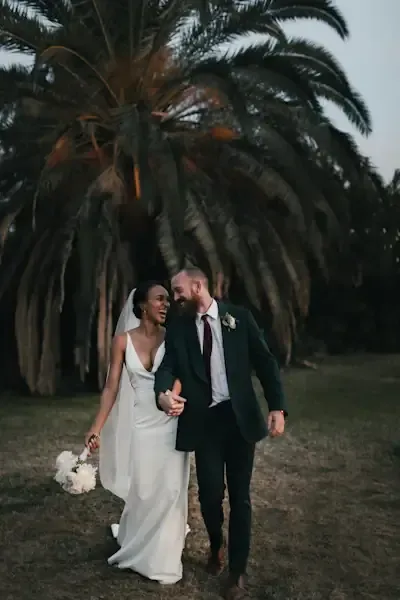 Priya and Marc laughing as they walk hand in hand beneath a palm tree at dusk