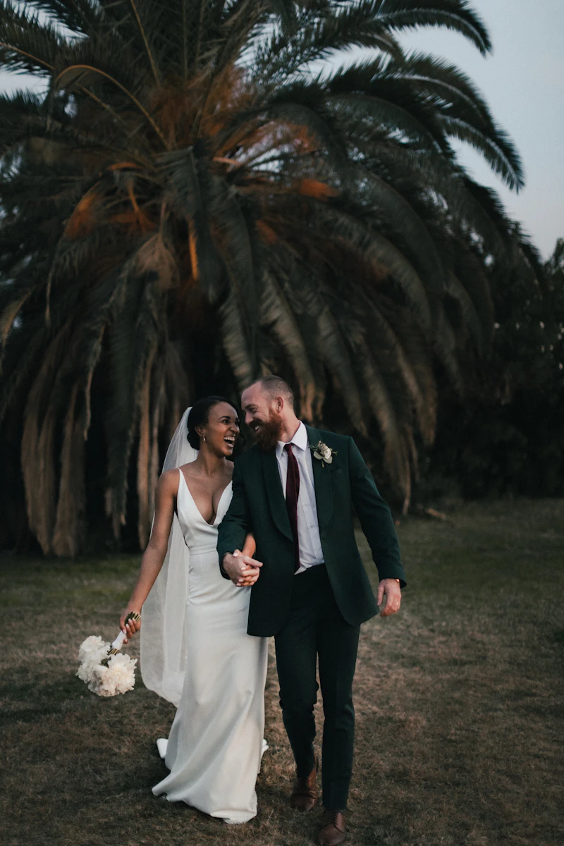 Couple in sleek wedding attire at twilight under palm trees, moody editorial lighting